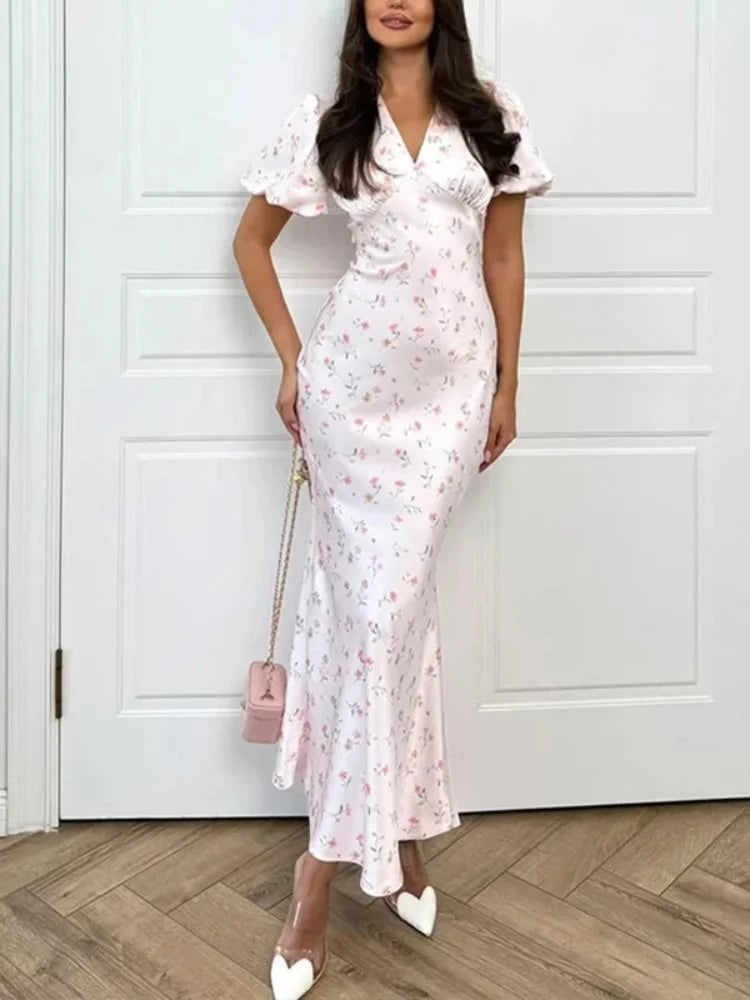 A woman models a white short-sleeve maxi dress with a pink and green floral print, featuring a V-neckline, ruffled sleeves, and a fitted silhouette. She is holding a small pink handbag and wearing white pointed-toe heels, standing in front of a white paneled door on a wood floor.