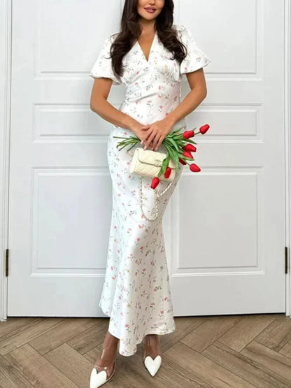 A woman models a white short-sleeve maxi dress with a delicate pink and green floral print, holding a small cream clutch and red tulips in front of a white paneled door.