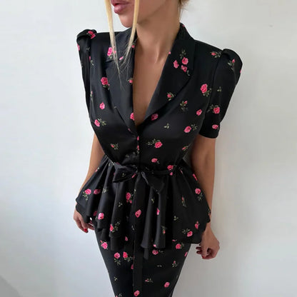 A woman wearing a black satin-like short-sleeve wrap dress with pink and green floral print, featuring puffed shoulders, a deep V-neckline, and a self-tie waist sash.