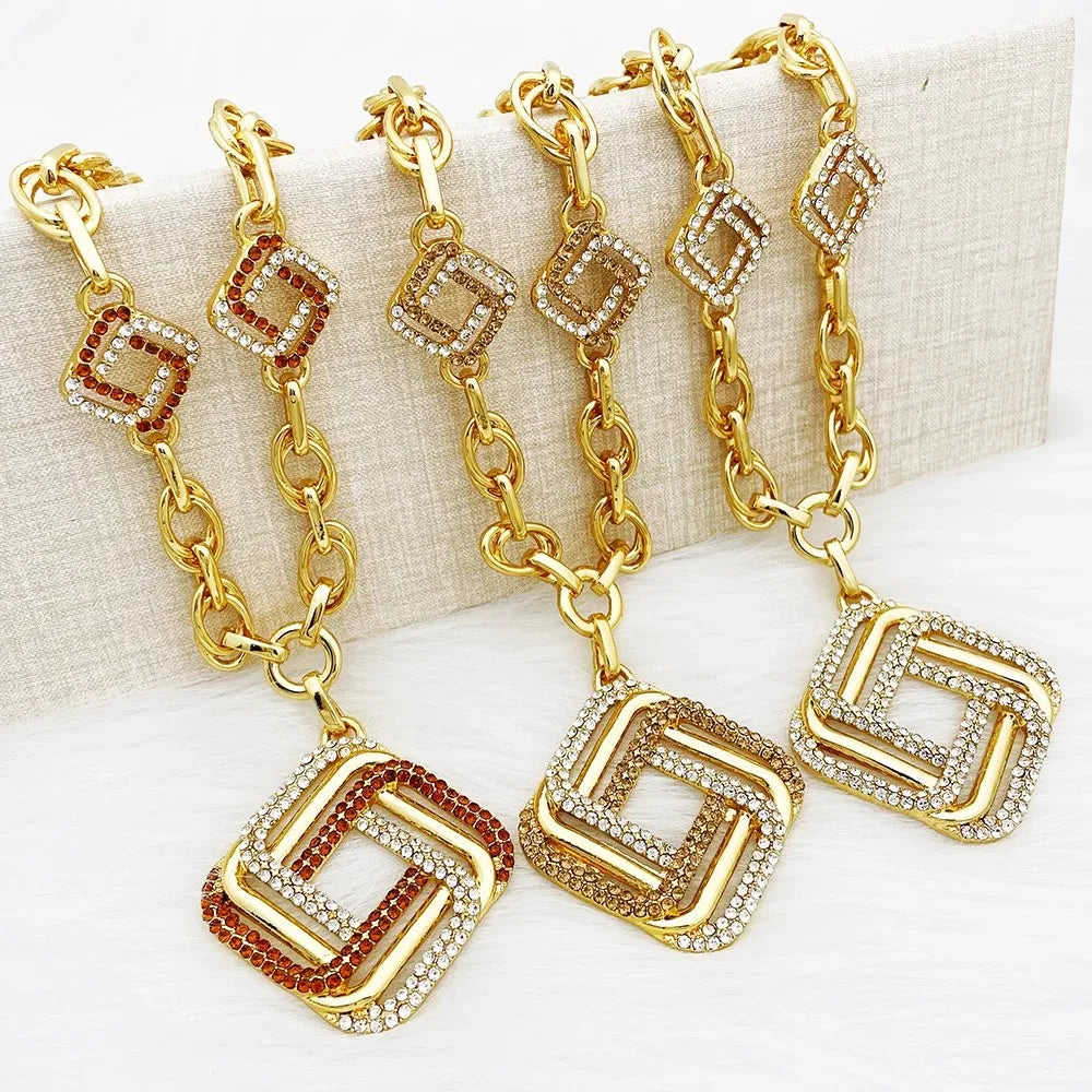 Four gold-tone chain keychains with geometric square charms, displayed on a textured beige block. The keychains feature clear rhinestones on some links and brown rhinestones on alternating sections of the chain and charms.