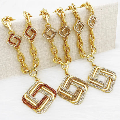 Four gold-tone chain keychains with geometric square charms, displayed on a textured beige block. The keychains feature clear rhinestones on some links and brown rhinestones on alternating sections of the chain and charms.