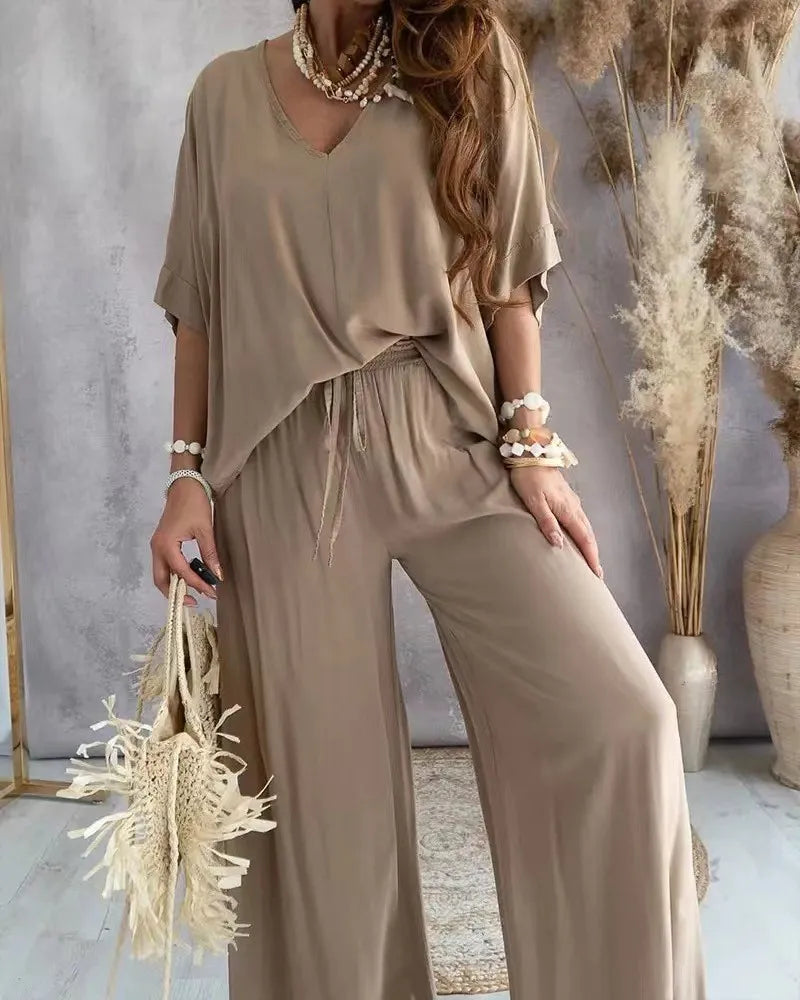 Woman wearing a beige outfit with wide-leg pants, holding dried plants against a neutral background.