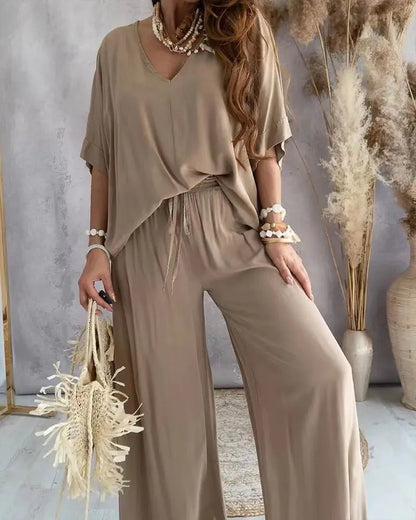 Woman wearing a beige outfit with wide-leg pants, holding dried plants against a neutral background.
