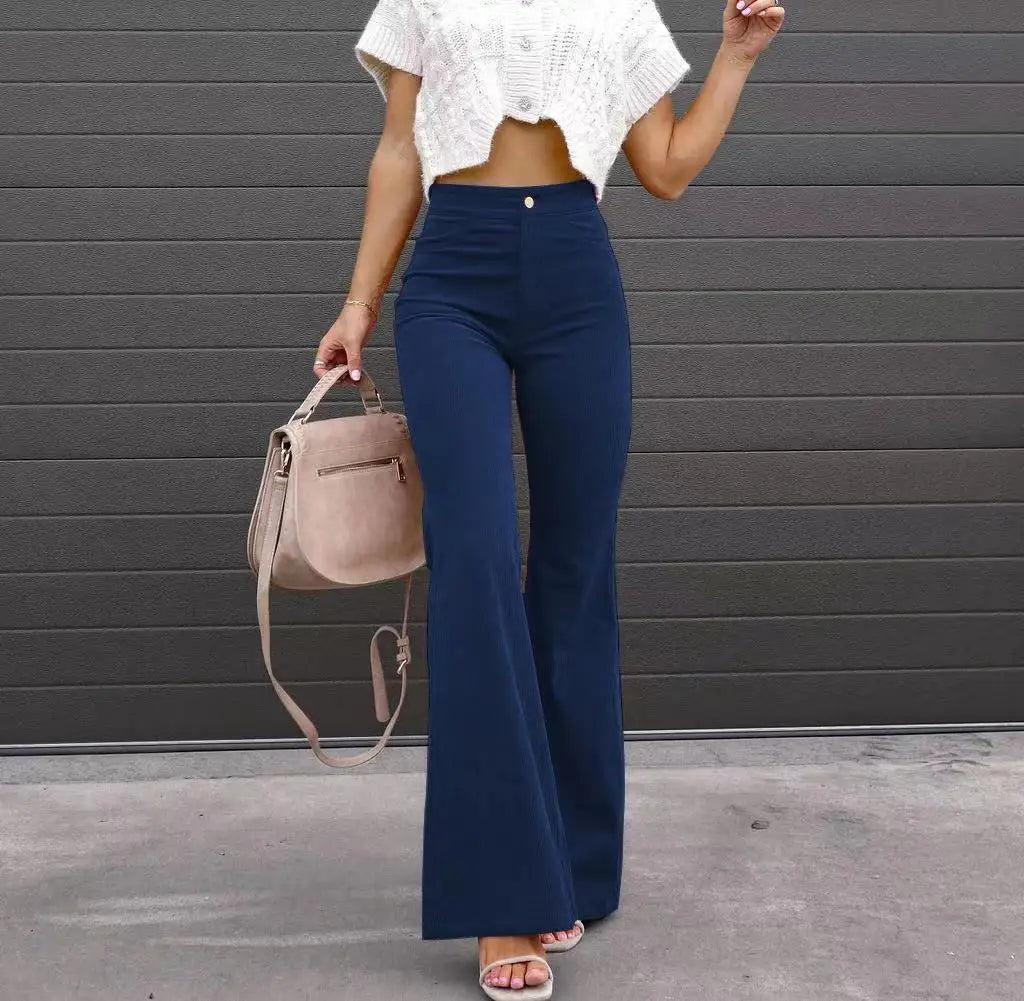 Person wearing a white top and blue high-waisted pants holding a beige handbag against a gray wall. Slim micro flare corduroy trousers with elastic waistband, screw thread button detail, casual style for women. Available in multiple colors UK FTF Market.