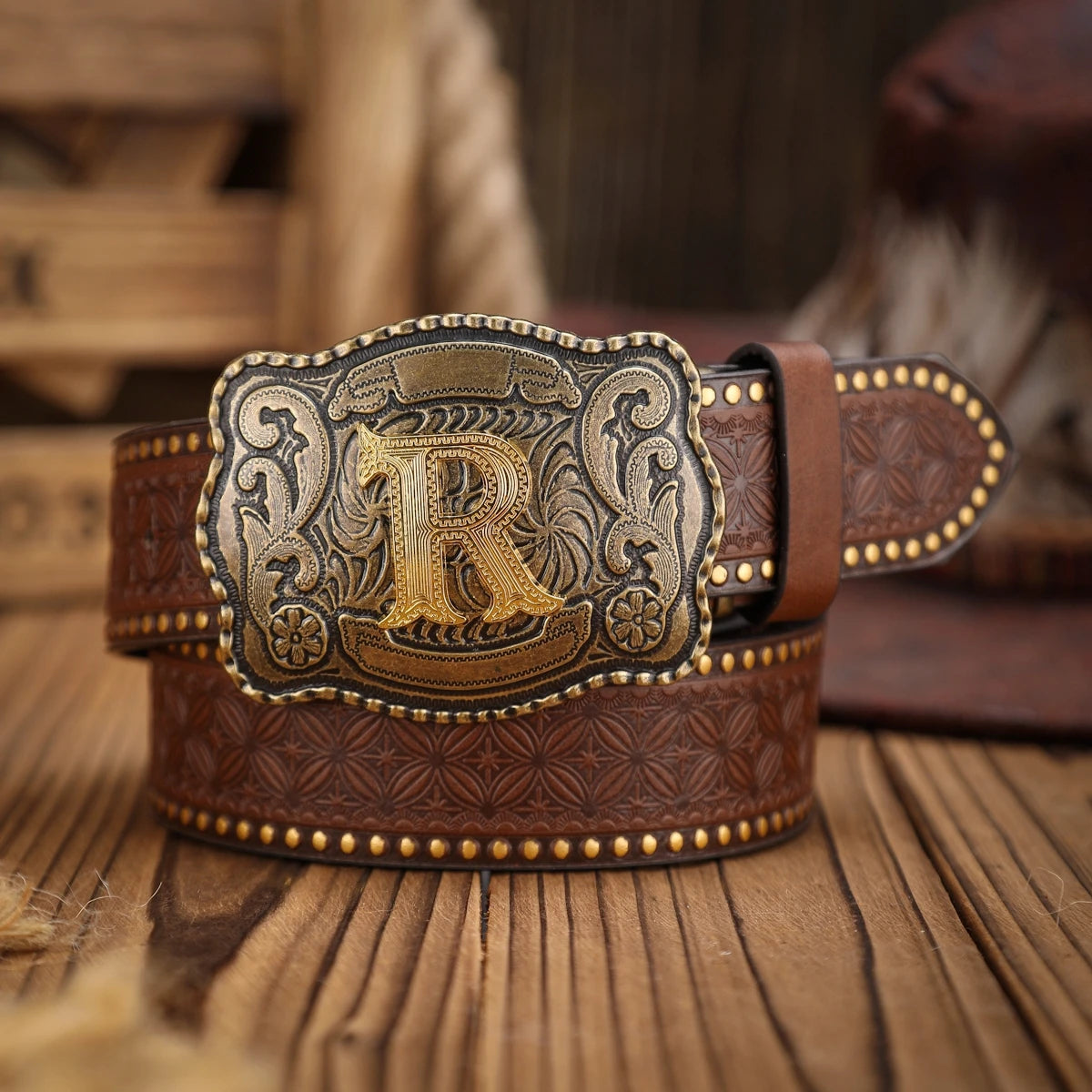Brown leather belt with gold and silver buckle featuring a large 'R' on a wooden surface.