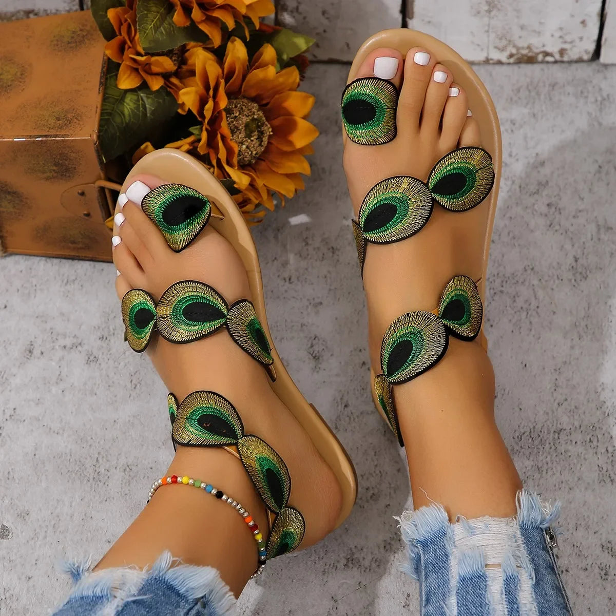 Beige sandals with peacock feather designs on a concrete floor.
