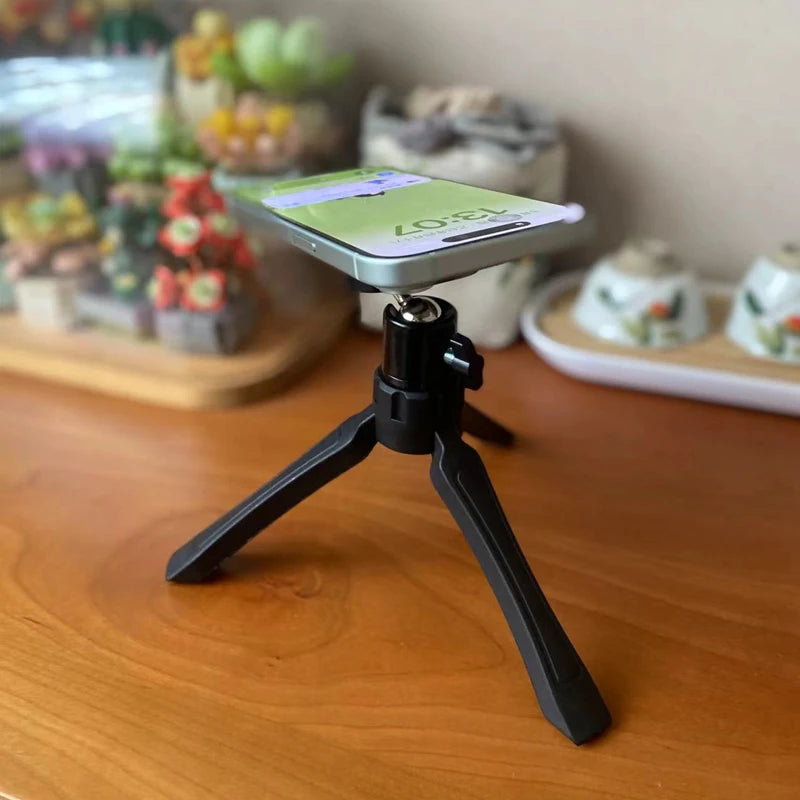 Small black tripod holding a smartphone on a wooden surface with blurred background. Magnetic phone tripod and wireless grip with remote control. Fits iPhone MagSafe and Samsung devices. Lightweight selfie stick tripod for creators.
