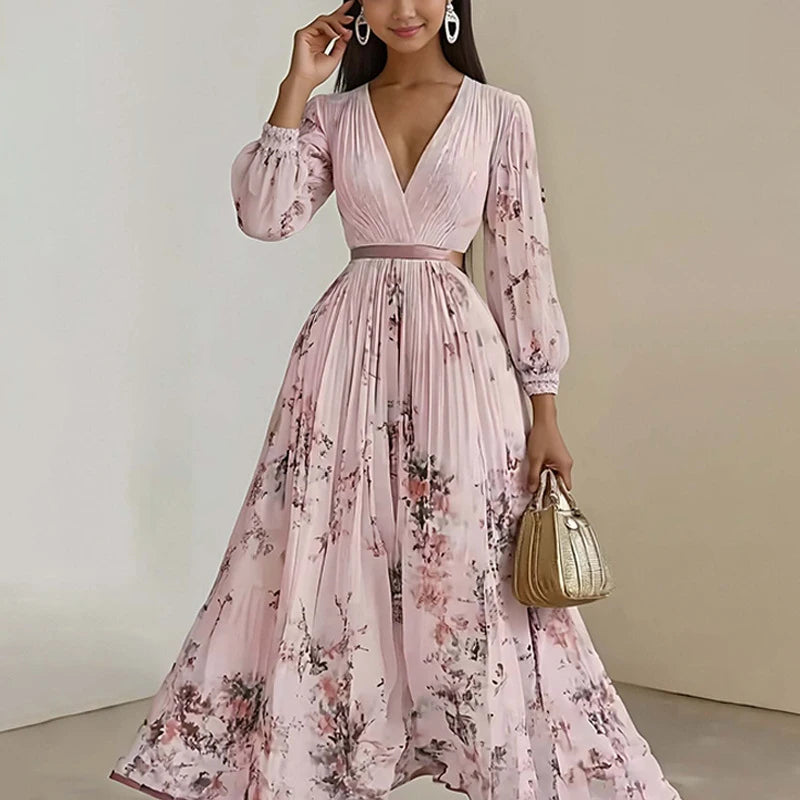 A woman models a long-sleeved, pleated maxi dress in light pink with a floral print. The dress features a V-neckline, a matching waistband, and a full skirt.