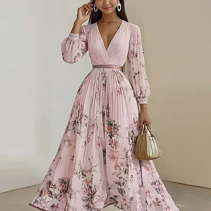 A woman models a long-sleeved, pleated maxi dress in light pink with a floral print. The dress features a V-neckline, a matching waistband, and a full skirt.