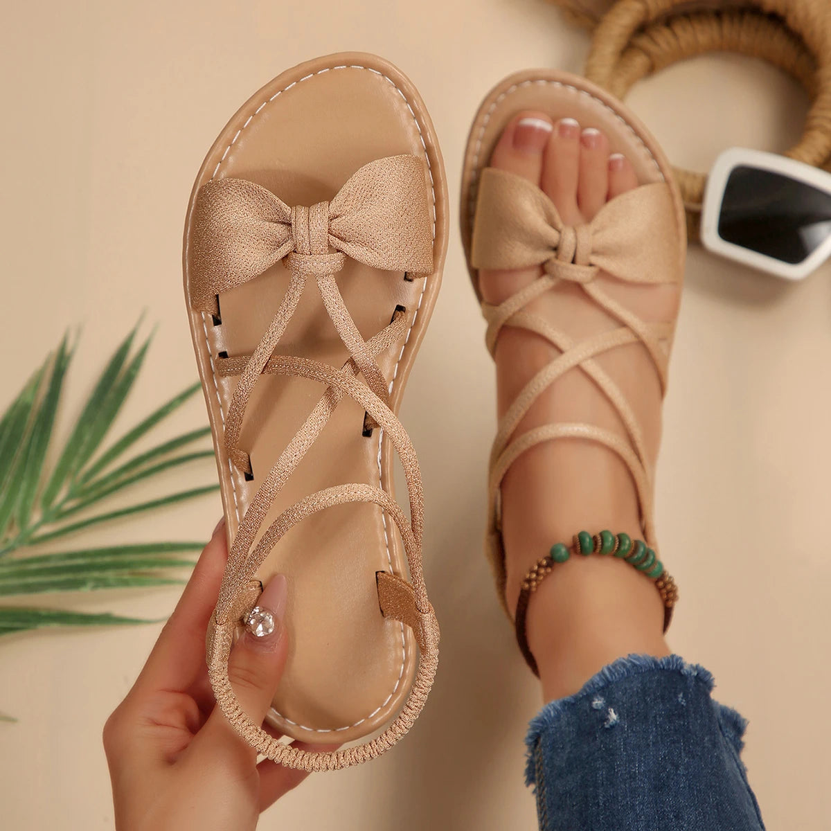 Beige sandals with bow detail held by a hand, on a neutral background.