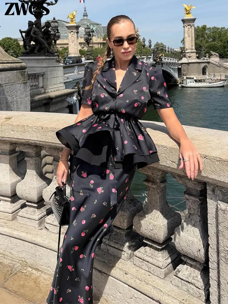 A woman models a black two-piece satin-like set with a pink floral print, featuring a short-sleeve collared top and matching wide-leg pants, posed on a stone bridge with water and historic architecture in the background.