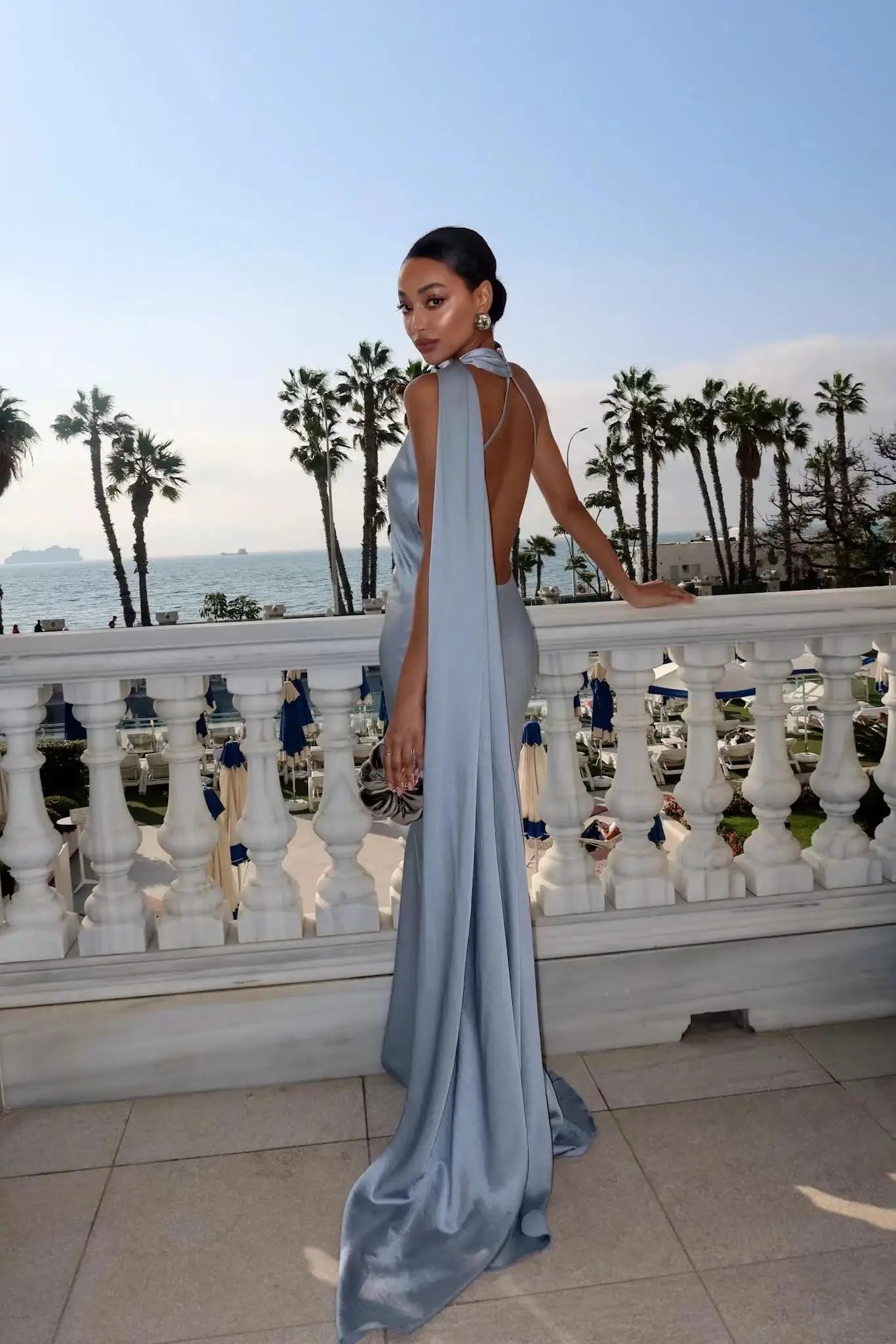 Woman in a light blue evening gown standing on a balcony with palm trees and ocean view.