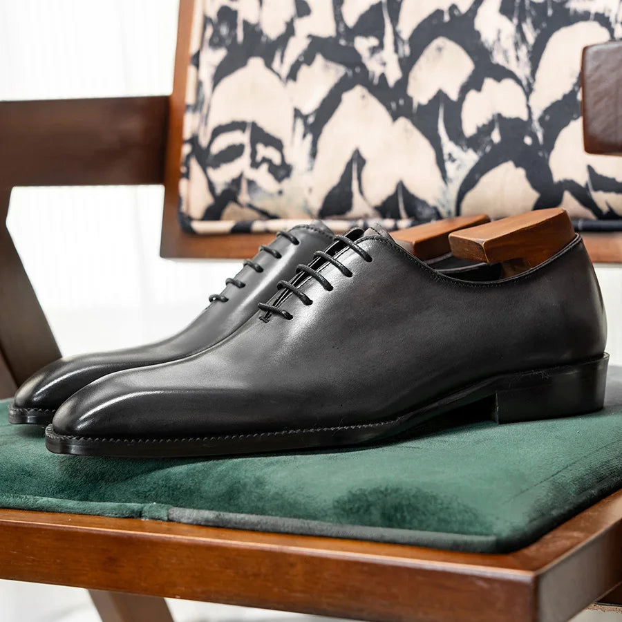 Black leather dress shoes on a green cushion with a patterned chair in the background