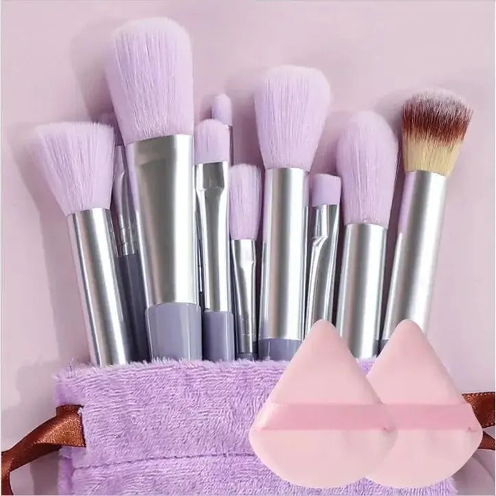 Makeup brushes with purple bristles and silver handles on a pink background, with two pink makeup sponges in front. Soft fluffy makeup brush set with wool fiber bristles and plastic handles. Complete set for powder, foundation, eyeshadow, and beauty blending.