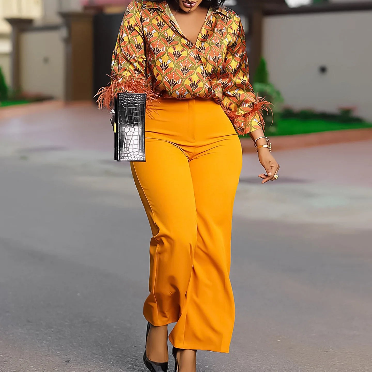 Woman in a patterned top and bright yellow pants walking outdoors. African print two-piece polyester outfit with satin printed shirt and high waist wide leg pants. Comfortable, elegant office wear for women.