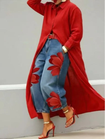 A model wearing a long red button-up shirt dress over blue denim pants with large red floral appliques, paired with red high-heeled sandals and gold jewelry, set against a plain gray wall.