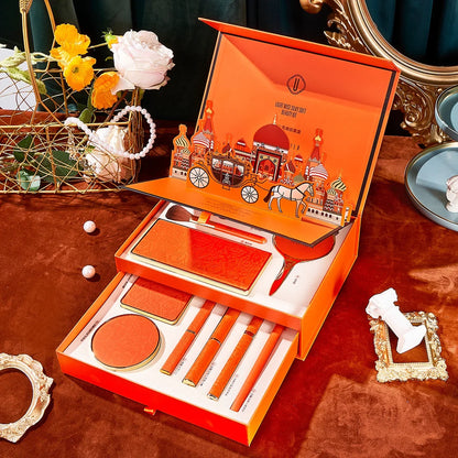 Orange makeup set with decorative packaging on a table with flowers and a mirror. Christmas makeup gift box with BB cream, lipstick, eyeshadow, eyebrow pencil, and brushes. Holiday beauty set for women & girls.