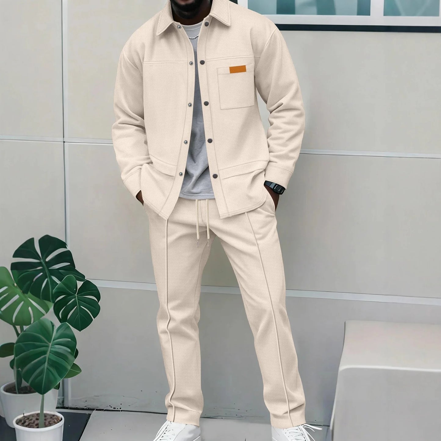 Person wearing a beige jacket and pants standing against a light gray wall with a plant to the left.