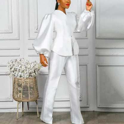 Woman in a white outfit standing in a room with a decorative wall and basket of flowers. Women's 2-piece pants set with solid satin blouse, straight high-waist trousers. Office lady luxury outfit for professional and elegant occasions.