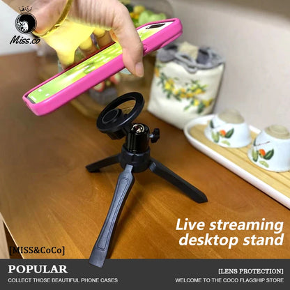 Hand holding a pink phone case with a live streaming desktop stand on a wooden surface. Magnetic phone tripod and wireless grip with remote control. Fits iPhone MagSafe and Samsung devices. Lightweight selfie stick tripod for creators.