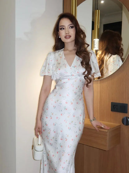 A woman wearing a white short-sleeve V-neck maxi dress with a delicate pink and green floral print, standing in front of a wooden vanity with a large round mirror.