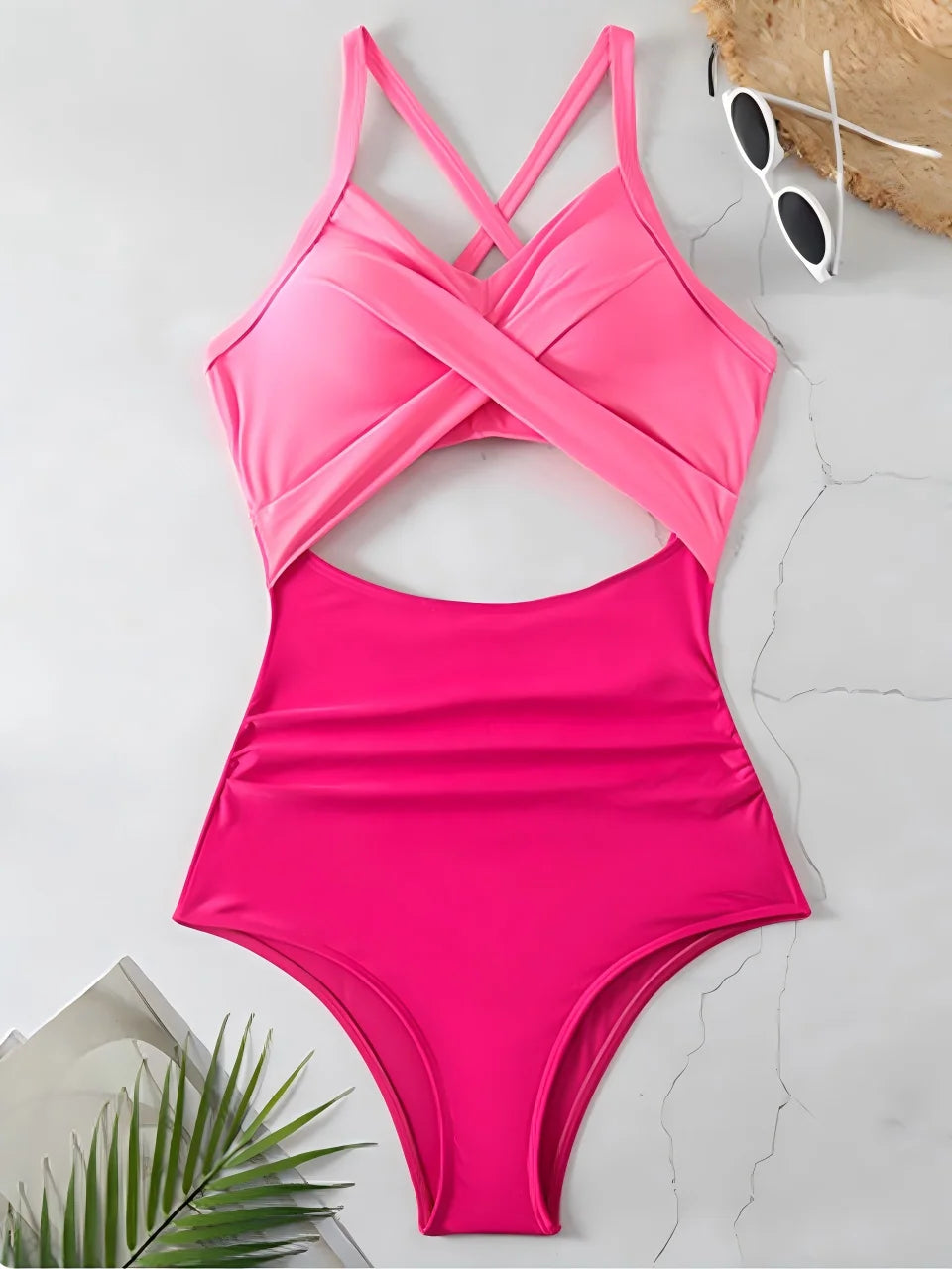 Pink one-piece swimsuit on a light gray surface with sunglasses and a palm leaf.