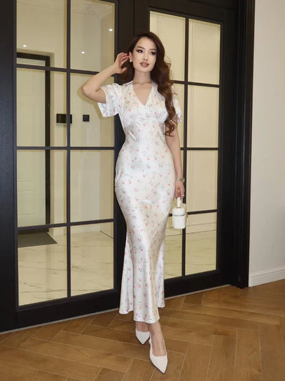 A woman models a white V-neck maxi dress with a delicate pink and green floral print, styled with white pointed-toe heels and a small white handbag, standing in front of a black-framed glass door.
