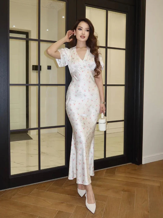 A woman models a white V-neck maxi dress with a delicate pink and green floral print, styled with white pointed-toe heels and a small white handbag, standing in front of a black-framed glass door.