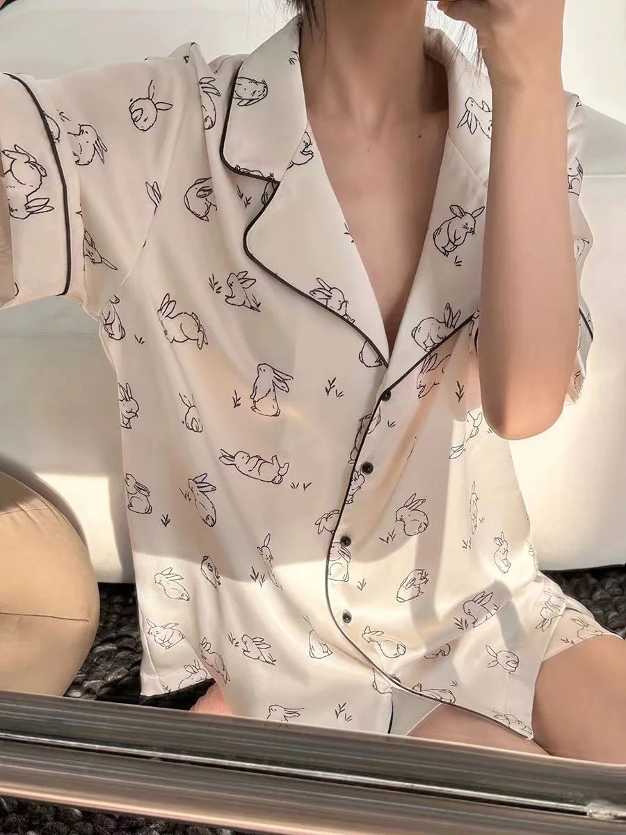 Person wearing a white pajama set with black patterns sitting on a chair. Women’s cherries print summer pajamas set—faux satin V-neck top and shorts, funny cute style. Lightweight, comfortable, UK fashion set.