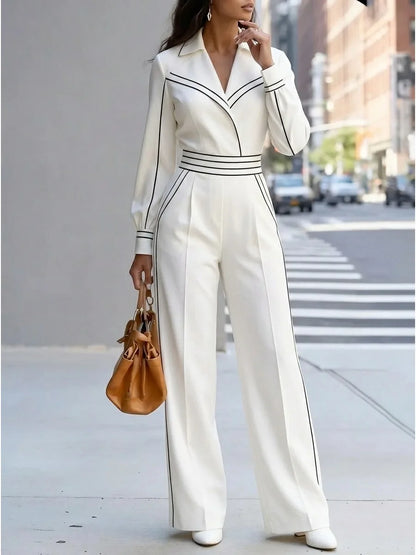 A woman stands on a city sidewalk wearing a white long-sleeve jumpsuit with thin black piping details and a matching belt. She is holding a brown leather handbag.