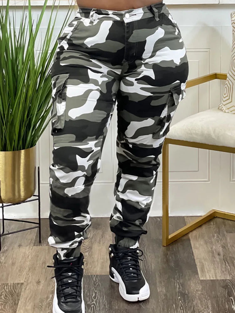 Person wearing camouflage pants and black sneakers in a room with a plant and chair.