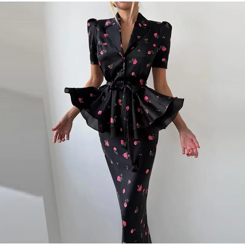 A woman modeling a black satin-like two-piece set with a pink and green floral print. The top is a short-sleeve blazer-style vest with puffed shoulders and a ruffled peplum hem, paired with a matching fitted skirt.