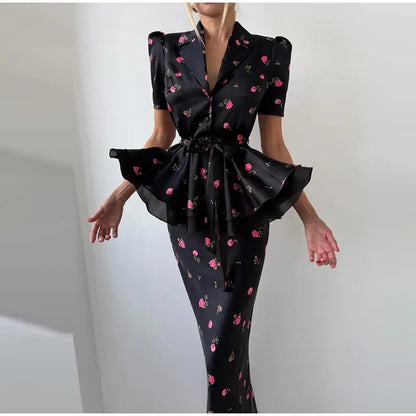 A woman modeling a black satin-like two-piece set with a pink and green floral print. The top is a short-sleeve blazer-style vest with puffed shoulders and a ruffled peplum hem, paired with a matching fitted skirt.