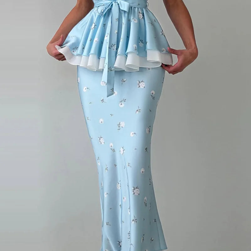 A light blue maxi dress with a ruffled peplum waist, tie detail, and a small white floral print, shown on a model against a neutral gray background.