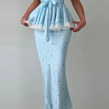 A light blue maxi dress with a ruffled peplum waist, tie detail, and a small white floral print, shown on a model against a neutral gray background.