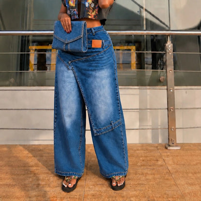 A person wearing wide-leg blue denim skirt pants with a diagonal front slit, holding a matching blue denim clutch bag, standing on a tiled floor in front of a glass railing.