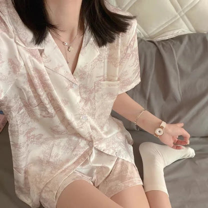 Woman wearing a light-colored satin robe sitting on a bed. Women’s cherries print summer pajamas set—faux satin V-neck top and shorts, funny cute style. Lightweight, comfortable, UK fashion set.