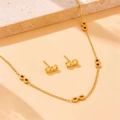 Gold jewelry set featuring a delicate chain necklace and matching stud earrings, both decorated with small oval links that have circular cutouts, arranged on a warm beige surface.