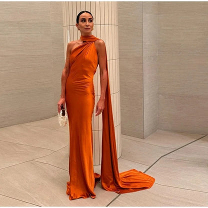 Woman in an orange evening gown with a long train standing against a neutral wall.