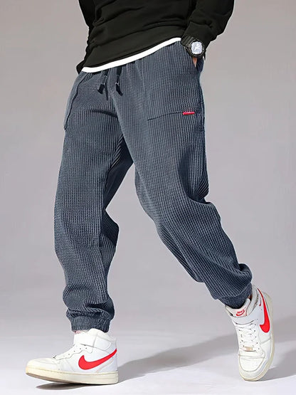 Person wearing gray checkered pants with a red tag, white sneakers, and a black jacket on a gray background. Men's baggy corduroy jogger pants, tech fleece sweatpants, autumn winter harem trousers, elastic waist, thermal, UK FTF Market.