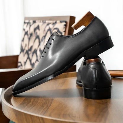 Black leather dress shoes on a wooden surface with a patterned chair in the background