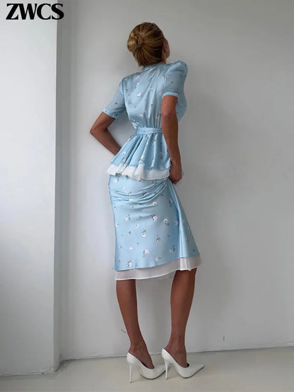 A woman stands with her back to the camera wearing a light blue short-sleeve midi dress with white ruffle trim and small white floral embroidery, paired with white pointed-toe heels. The brand name 'ZWCS' is visible in the top left corner.