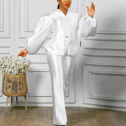 Person wearing a white outfit in a room with decorative walls and a basket of flowers. Women's 2-piece pants set with solid satin blouse, straight high-waist trousers. Office lady luxury outfit for professional and elegant occasions.