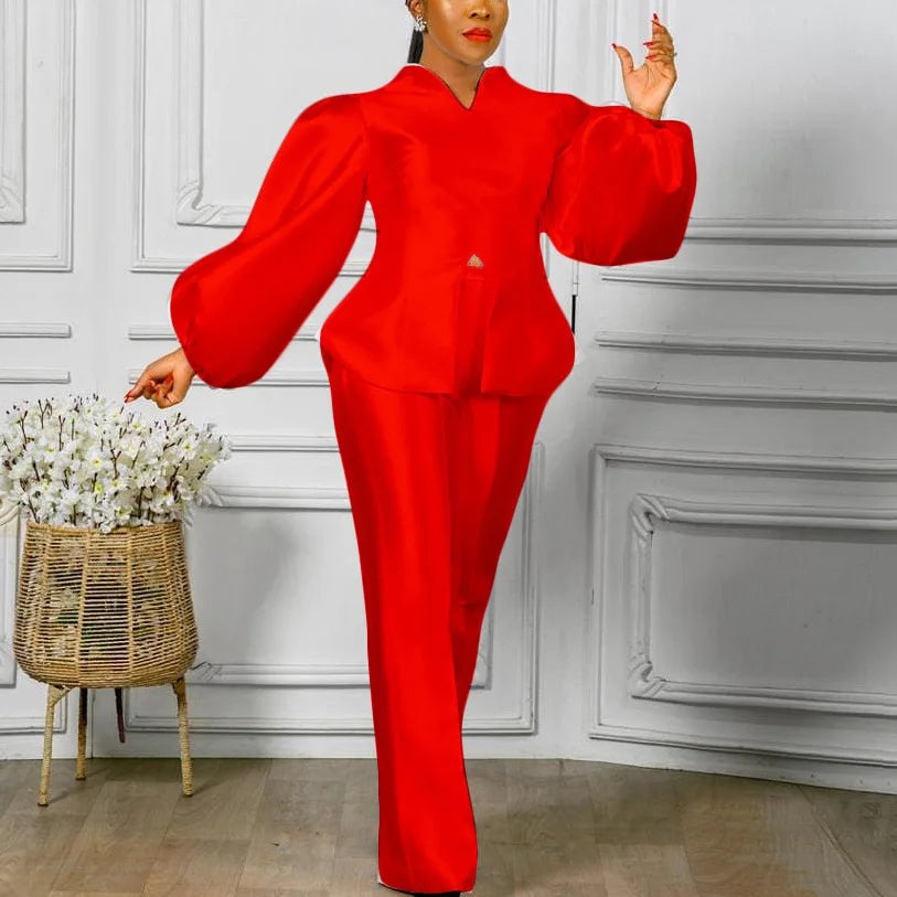 Person wearing a red outfit standing in a room with decorative white walls and a woven basket. Women's 2-piece pants set with solid satin blouse, straight high-waist trousers. Office lady luxury outfit for professional and elegant occasions.