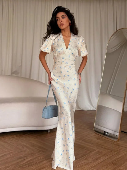 A woman models a cream-colored, floor-length dress with a delicate blue floral print. The dress features short puff sleeves, a V-neckline, and a fitted silhouette.