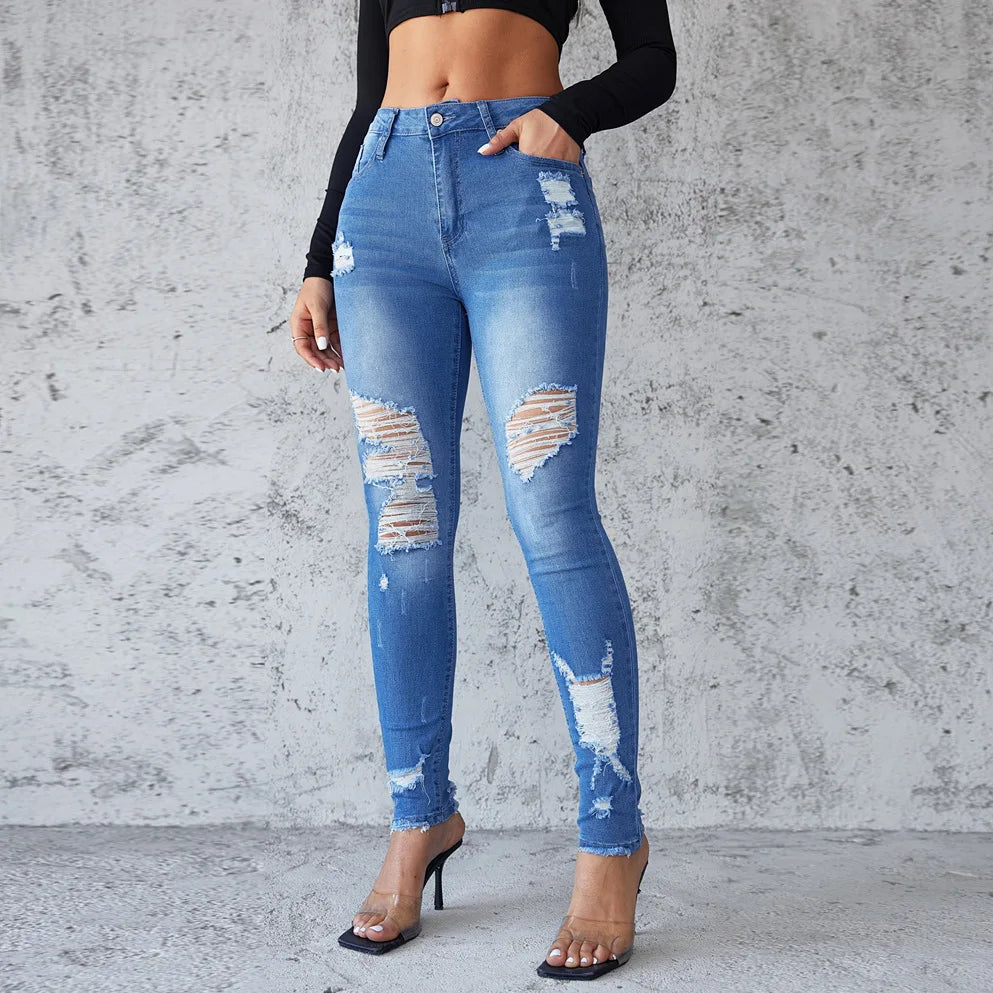 Person wearing blue distressed jeans against a gray concrete wall. Women’s high waist ankle length skinny jeans, ripped and distressed, pockets, solid slim pencil fit. Vintage streetwear by ftf fashion UK.
