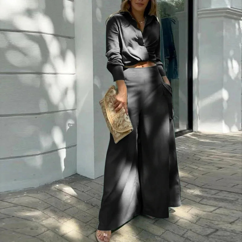Woman in a black outfit with wide-leg pants holding a clutch, standing on a sunlit pavement. Women's elegant pants set with long sleeve crop shirt and high waist wide leg trousers. Office lady style. Solid, flattering, comfortable fit.