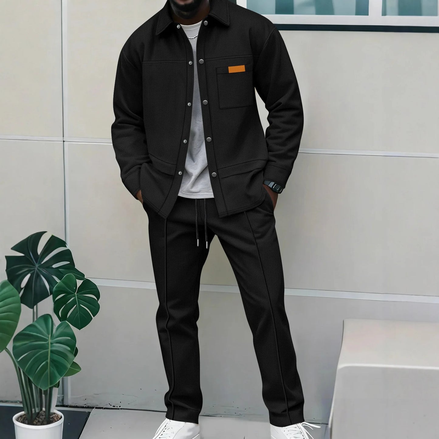 Person wearing a black jacket and pants standing outdoors with a plant and bench in the background.