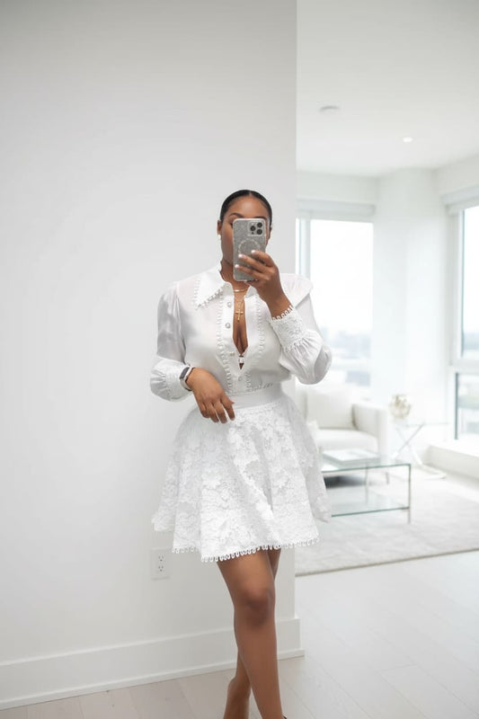 A woman takes a mirror selfie in a bright, modern white room, wearing a white long-sleeve blouse with a deep plunging neckline and a matching white lace mini skirt.