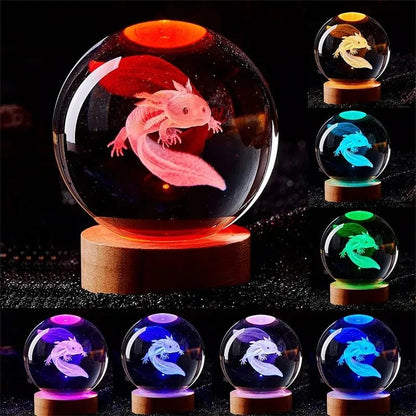Glowing crystal ball with axolotl design on a wooden base, surrounded by other crystal balls with different designs. 3D laser engraved axolotl crystal ball lamp with USB LED base, vibrant colours UK.